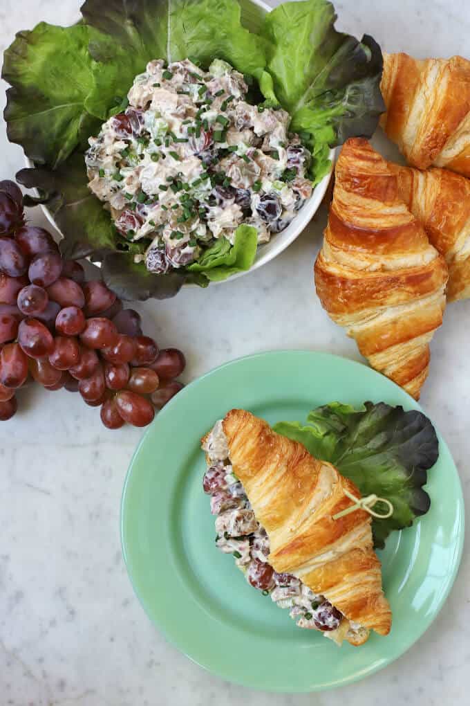 A flat lay on a white marble background containing chicken salad, grapes, and croissants with one filled croissant on a green plate.