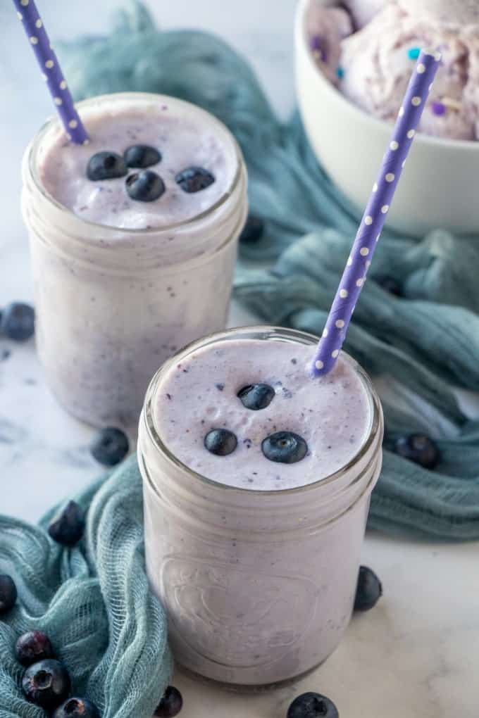 Blueberry milkshakes in jars with blueberries on top and purple straws.