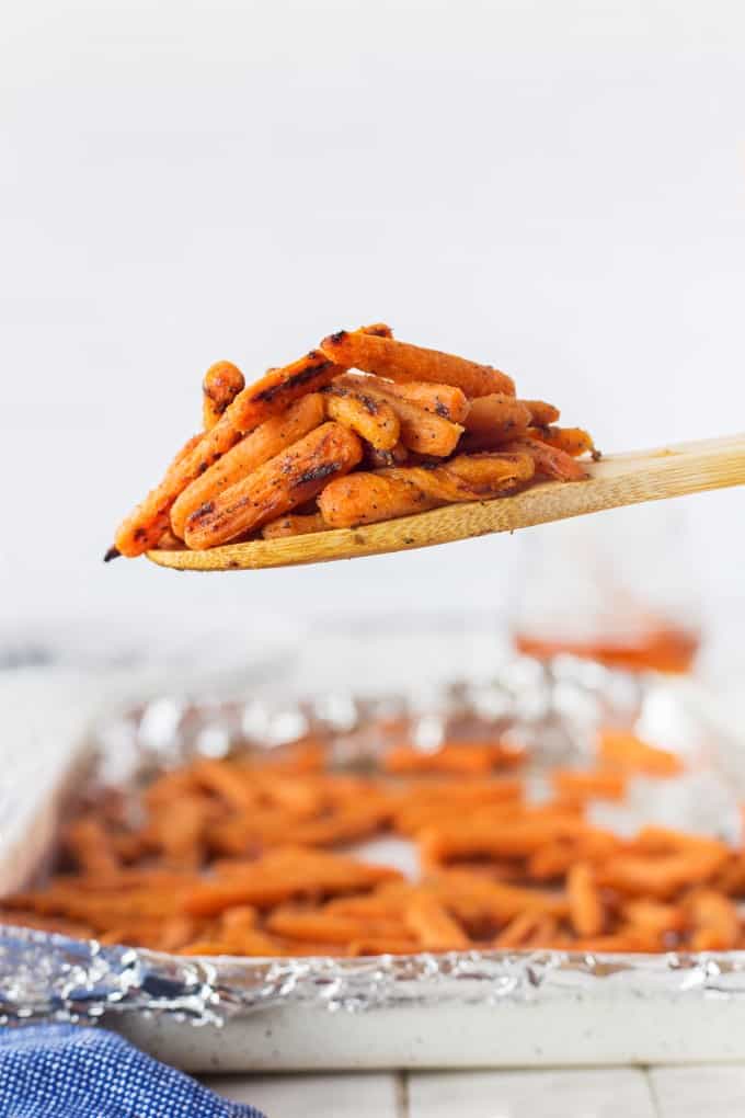 A wooden spoonful of roasted carrots with a baking sheet of carrots in the background