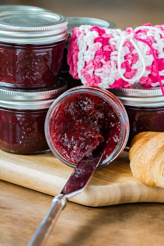 A knife into a jar of jam on its side with jars of jam, dishcloth and croissant. 