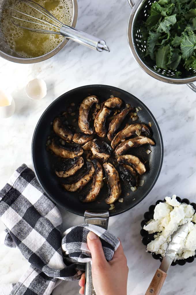 Top view of a cooked mushrooms organized in a spiral in a pan with a hand holding the handle with a dish towel, some fresh kale on the side. 