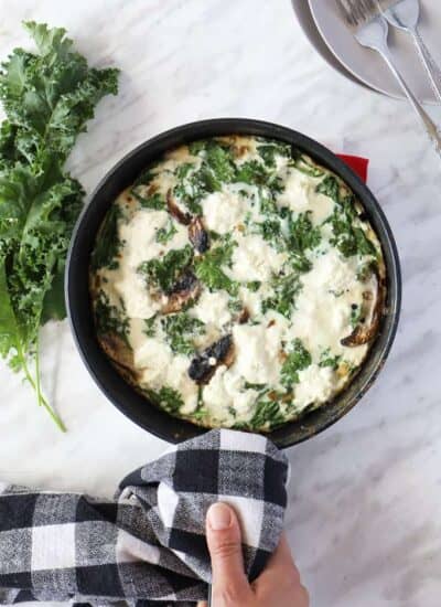 Top view of a cooked frittata in a pan with a hand holding the handle with a dish towel, some fresh kale on the side.