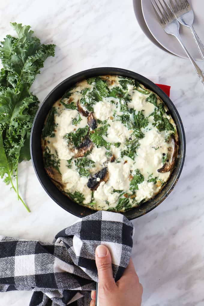 Top view of a cooked frittata in a pan with a hand holding the handle with a dish towel, some fresh kale on the side.
