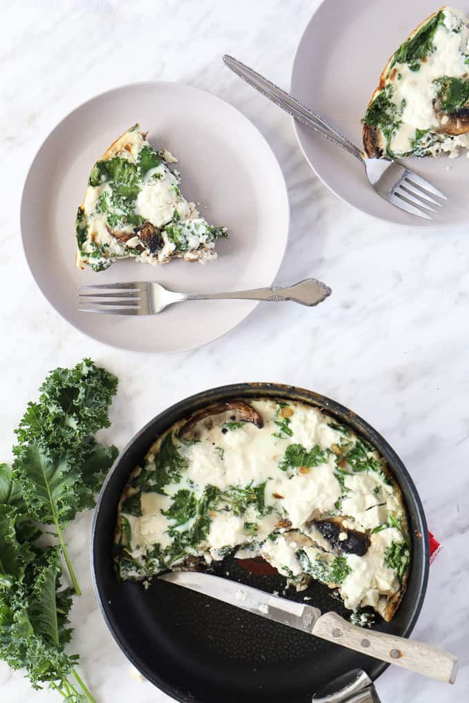Top view of a cooked frittata sliced in 2 plates with forks and the rest in the pan, some fresh kale on the side. 