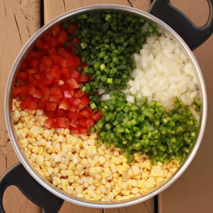 A large pot holding yellow corn and finely diced red peppers, jalapenos, onions, and garlic scapes.
