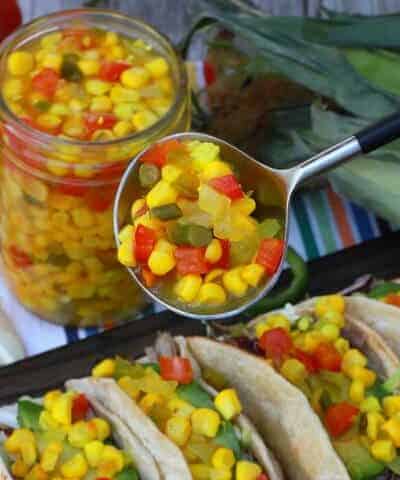 A spoon holds brightly coloured corn, red peppers, and jalapeno relish above tacos.