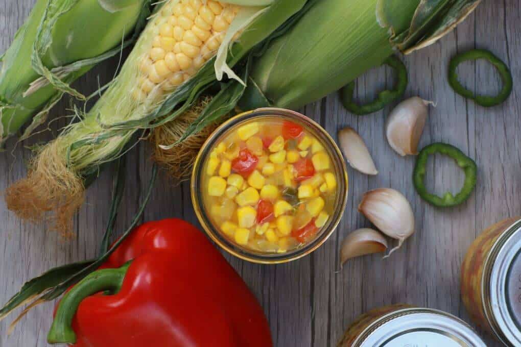 A jar filled with bright yellow corn, red peppers, and green jalapenos in the centre of fresh ingredients.