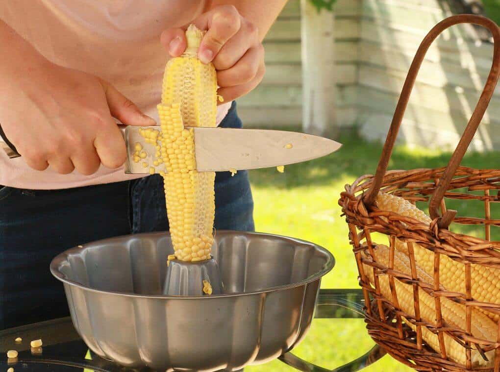 A person holds a cob of corn in the centre of a bundt pan while cutting the kernels off with a knife. 