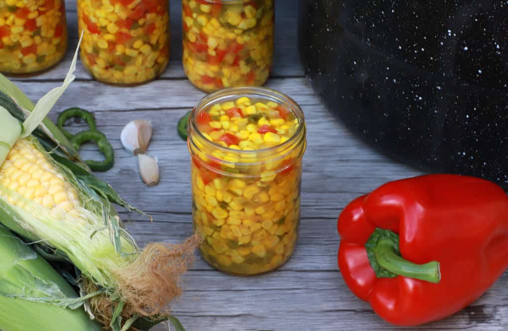A jar filled with bright yellow corn, red peppers, and green jalapenos in the centre of fresh ingredients and more filled jars.