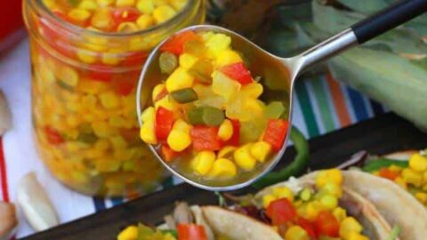 A spoon holds brightly coloured corn, red peppers, and jalapeno relish above tacos.