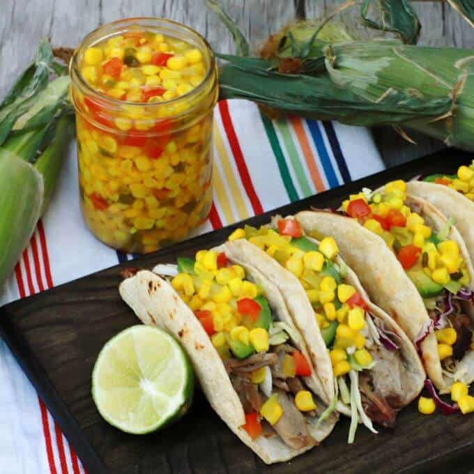 Three pork tacos topped with corn relish with a jar of bright yellow corn relish in the background.