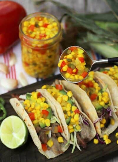 Three pork tacos topped with corn relish with a jar of bright yellow corn relish in the background.