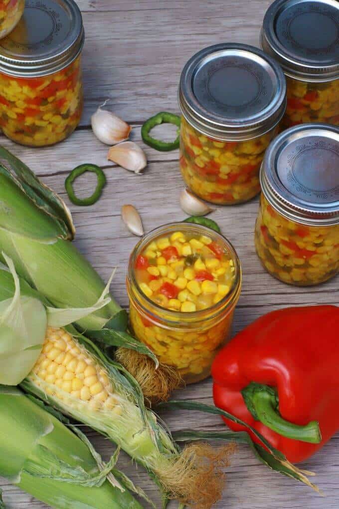 A jar filled with bright yellow corn, red peppers, and green jalapenos in the centre of fresh ingredients and more filled jars with lids..
