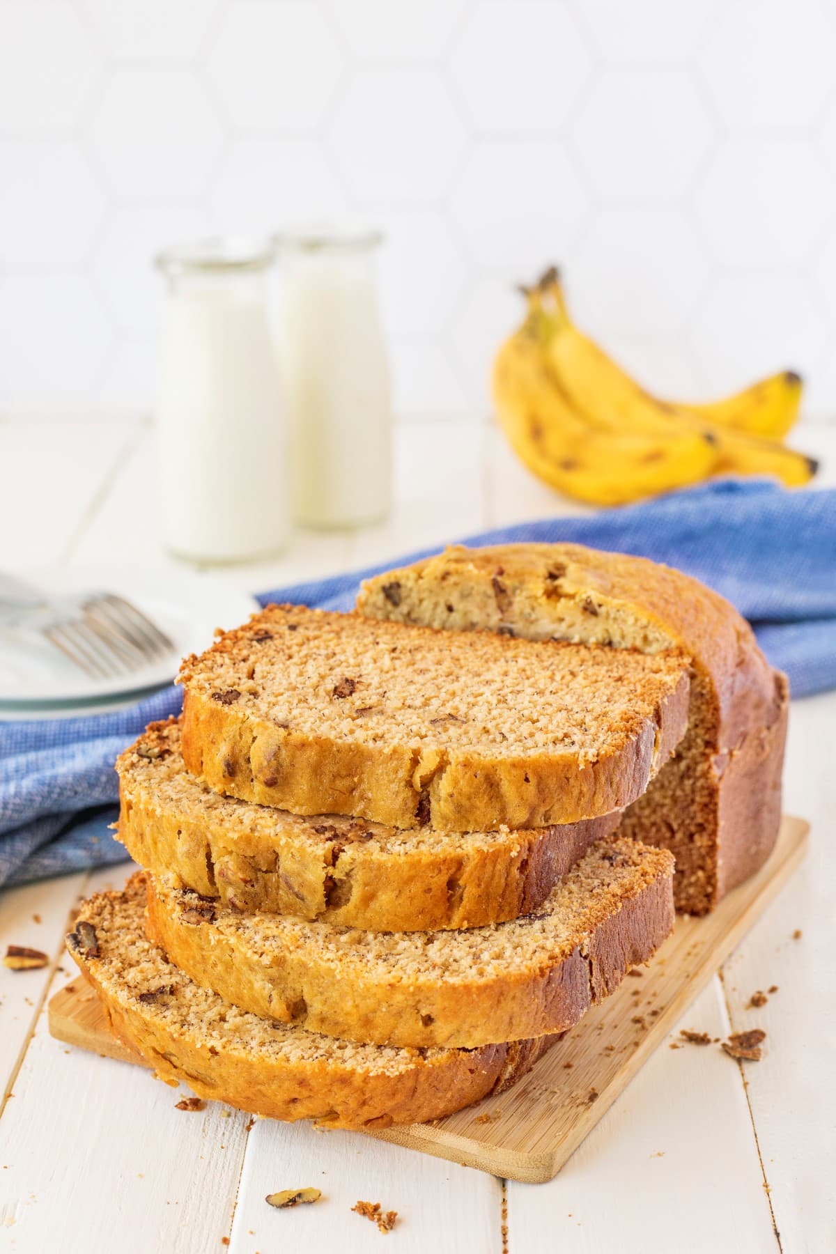 Stacked banana bread on a wooden board with milk and bananas in the background. 