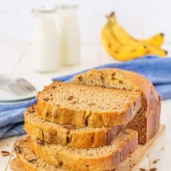 Stacked banana bread on a wooden board with milk and bananas in the background.