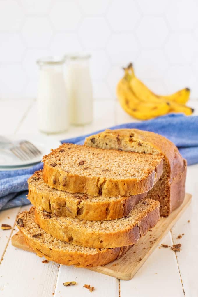 Stacked banana bread on a wooden board with milk and bananas in the background.