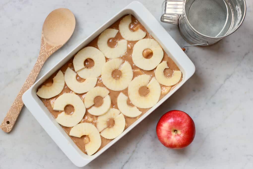 A white baking pan holds an unbaked Apple Dapple Cake topped with slices of apple.
