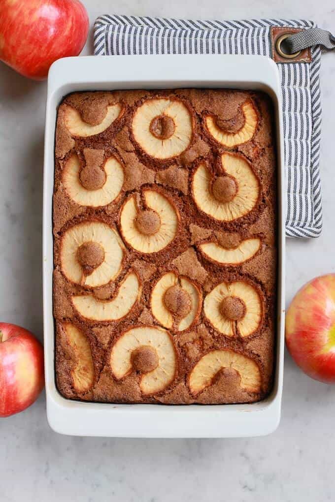 A white baking pan holds a golden baked Apple Dapple Cake topped with slices of apple.