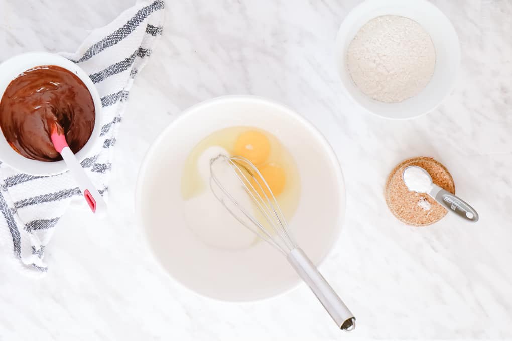 Top view of ingredients, eggs and sugar in a big bowl with a whisk, a smaller bowl on the left with melted chocolate and a bowl of flour on the right.