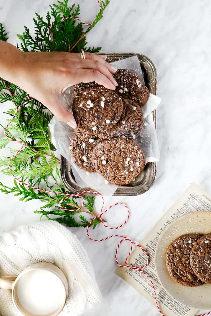 Overhead shot of brownie cookies with a hand grabbing one and greenery and white and red thread. 