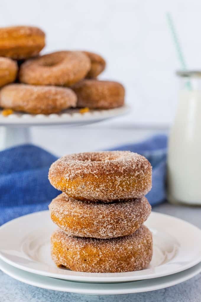 Stack of three donuts on a white plate with donuts in the background on a cake stand. 