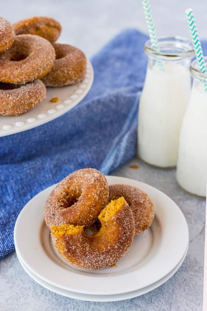 Pumpkin Donuts on a white plate, one broken in half. Milk in glass milk bottles in the background and donuts on a cake stand. 