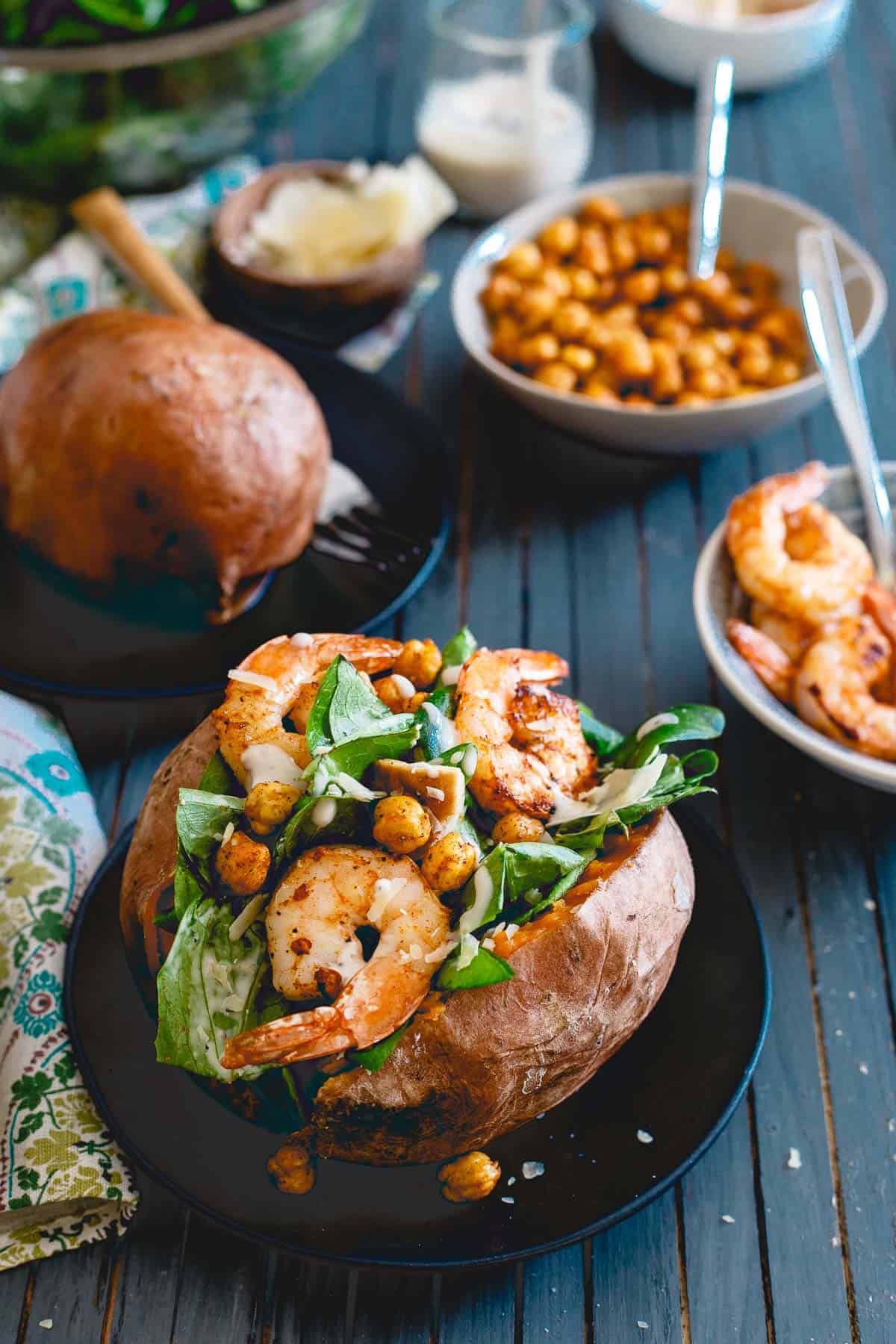Caesar Salad Stuffed Sweet Potatoes With Spicy Shrimp on a small black plate with a bowl of shrimp and chickpeas beside it.