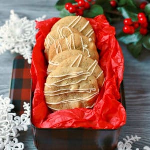 Golden baked cookies with white drizzle in a red tissue paper lined cookie tin surrounded by snowflakes.
