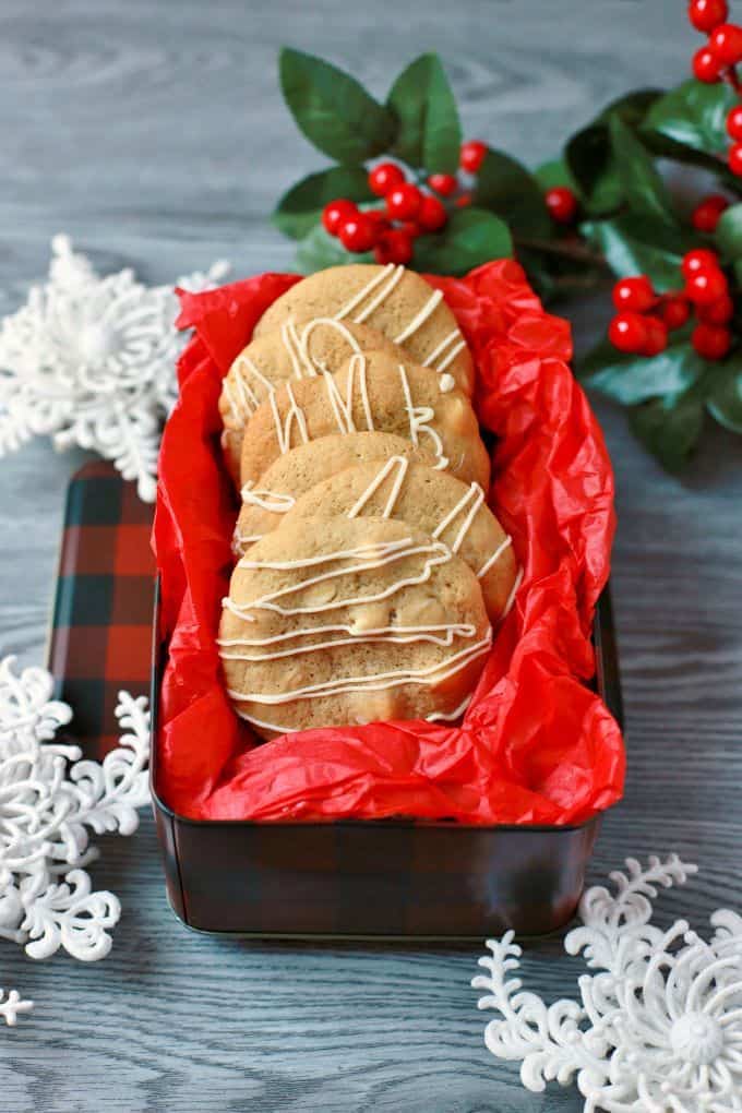 Golden baked cookies with white drizzle in a red tissue paper lined cookie tin surrounded by snowflakes.