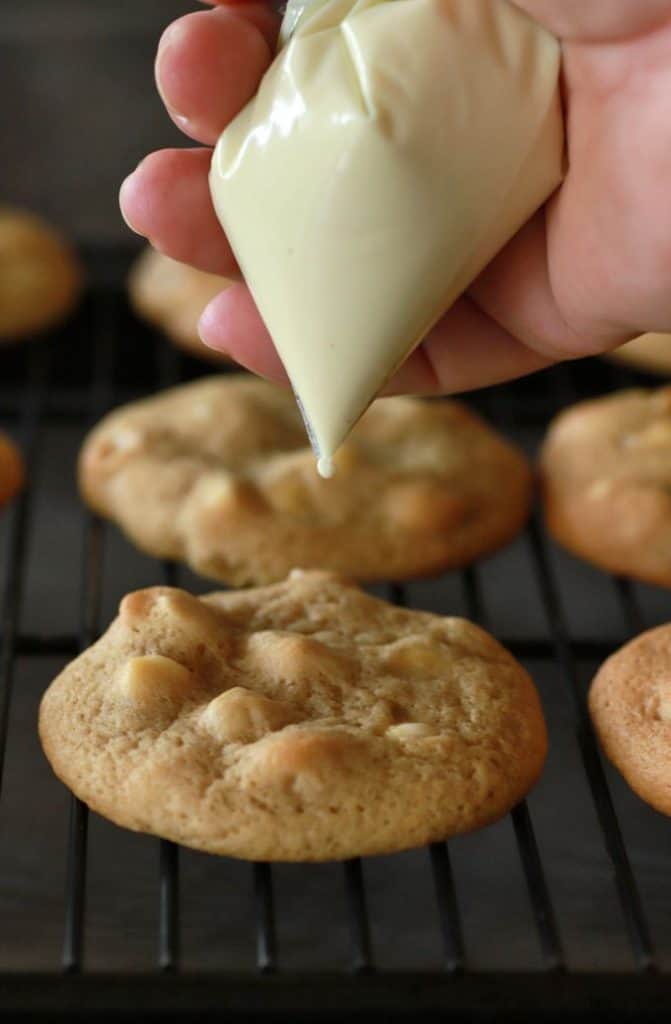 A golden baked cookie with a piping bag of melted white chocolate held over, ready to drizzle.