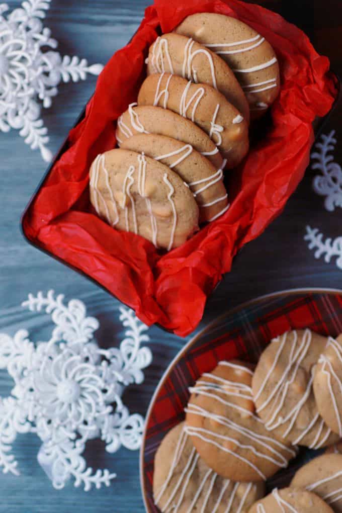 Golden baked cookies with white drizzle in a red tissue paper lined cookie tin surrounded by snowflakes.