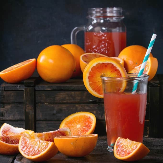 Sliced and whole Sicilian Blood oranges and glass of fresh red orange juice over old wooden table. Dark rustic style. Square image