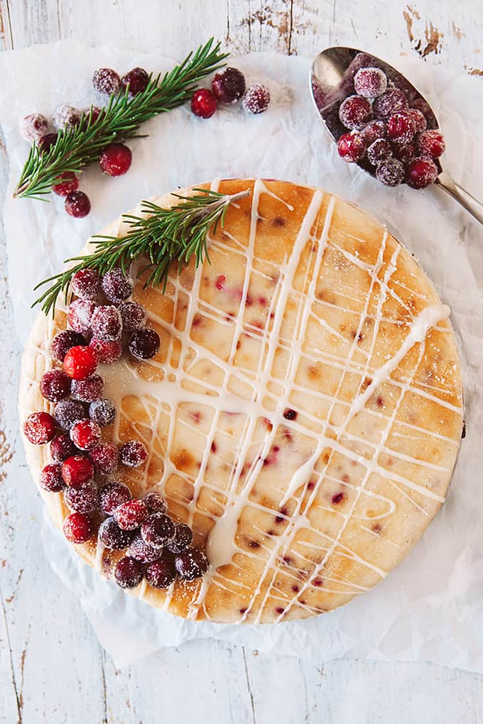 Cranberry White Chocolate Cheesecake on parchment paper on a white board decorated with cranberries and rosemary. 