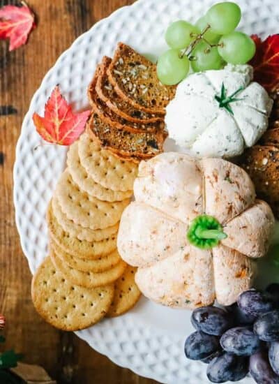 Top view of 2 pumpkin cheese ball with crackers and grapes on a white plate.