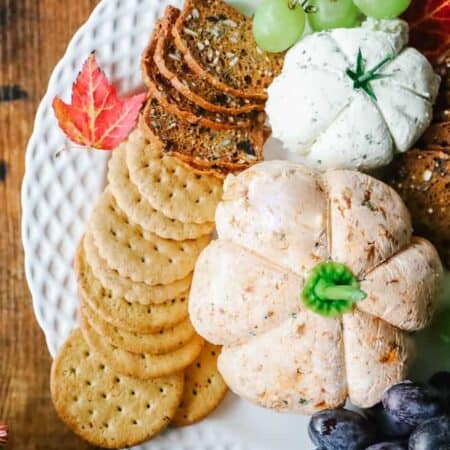 Top view of 2 pumpkin cheese ball with crackers and grapes on a white plate.