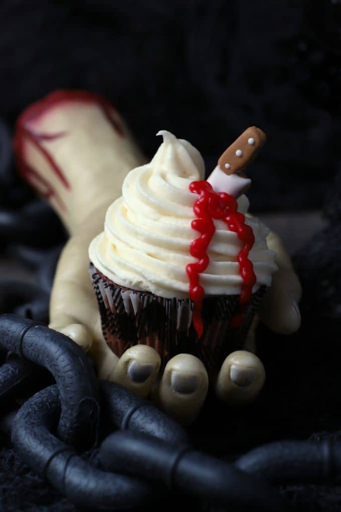 A bloody hand holds a cupcake topped with swirled white frosting oozing blood from a candy knife.