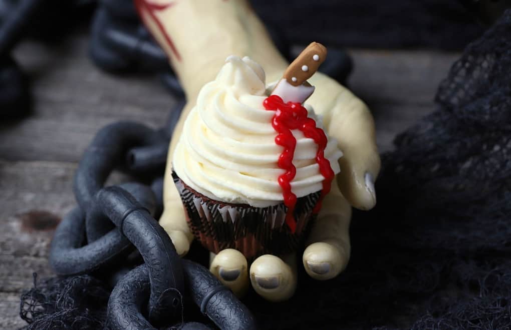 A bloody hand holds a cupcake topped with swirled white frosting oozing blood from a candy knife.