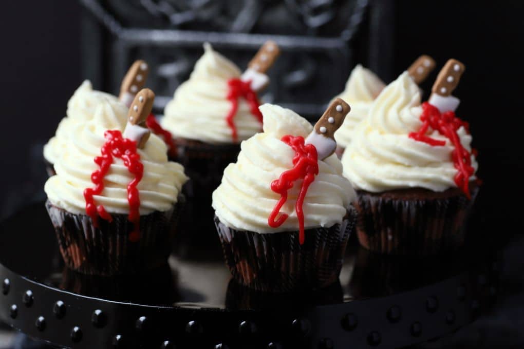 A platter of cupcakes topped with swirled white frosting oozing blood from a candy knife.