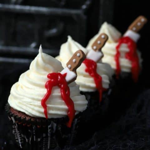 Three cupcakes topped with swirled white frosting oozing blood from a candy knife.