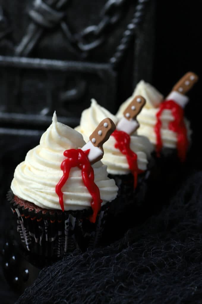 Three cupcakes topped with swirled white frosting oozing blood from a candy knife.