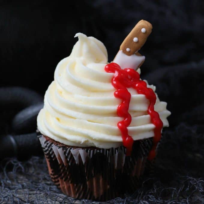 A single cupcake topped with swirled white frosting oozing blood from a candy knife.