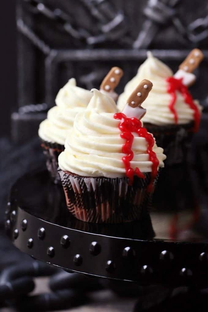 Three cupcakes topped with swirled white frosting oozing blood from a candy knife.
