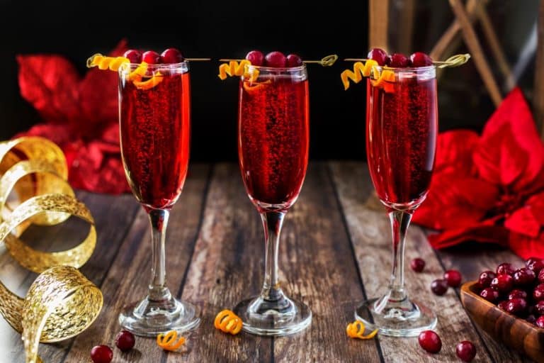 Three glasses of red sparkling drinks are garnished with cranberries and orange twists on skewers. The glasses are set on a rustic wooden table with red flowers, gold ribbon, and scattered cranberries in the background.