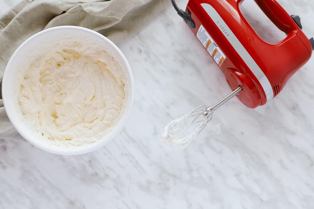 Whipped cream in a bowl with a red hand mixer lying down beside. 