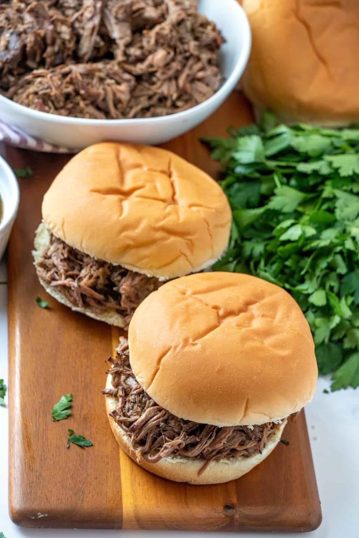 Two slow cooker beef dip sandwiches on a board with a bowl of pulled beef in the background. 