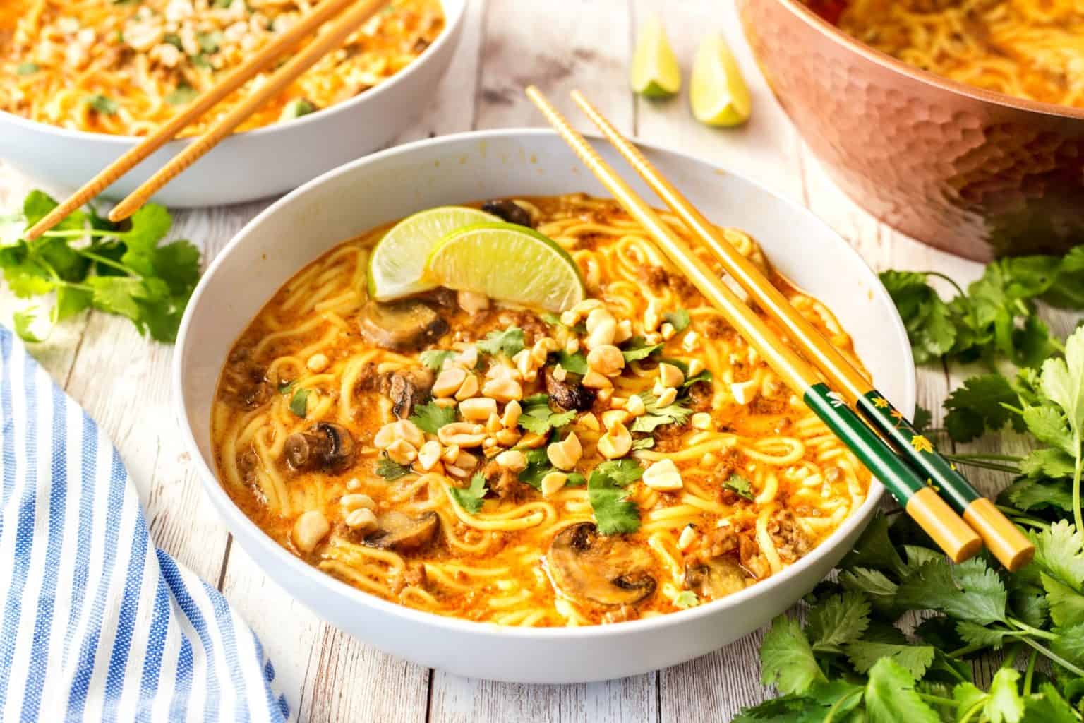 A bowl of noodle soup with creamy broth, topped with lime wedges, chopped peanuts, mushrooms, and fresh herbs. Chopsticks rest on the bowl's edge. Additional bowls and ingredients are visible in the background on a wooden surface.