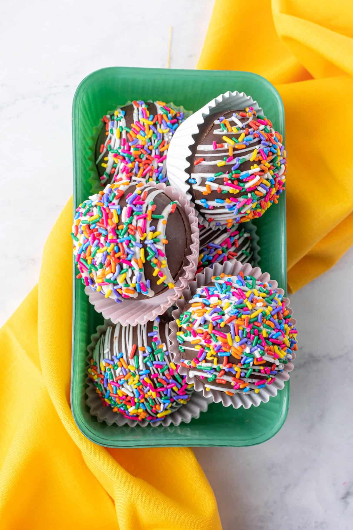 Overhead shot of Funfetti Strawberry Hot Chocolate Bombs in a green container with a yellow napkin. 