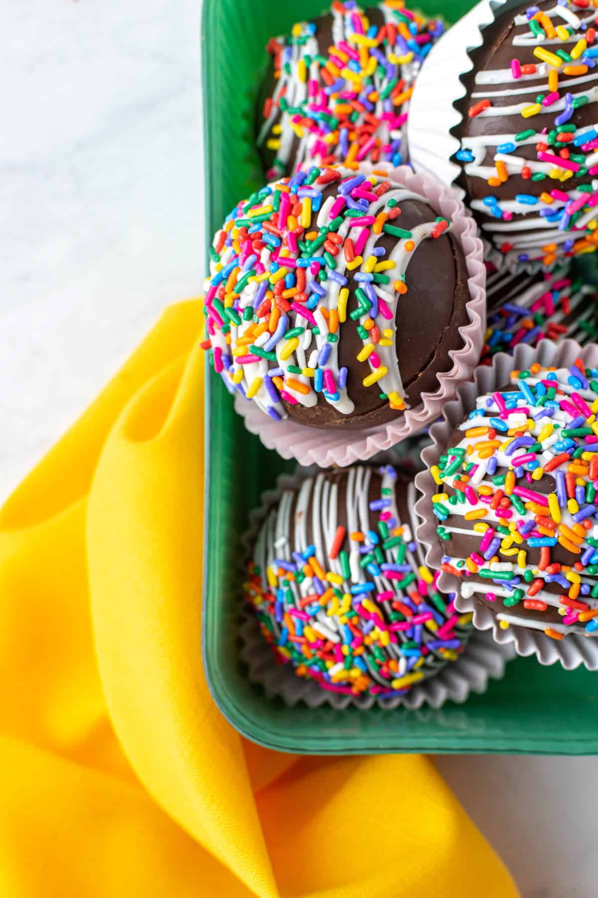 Overhead shot of hot cocoa bombs in a green container with a yellow napkin. 