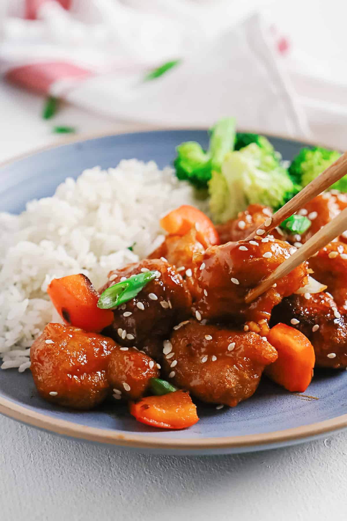 Picking up sweet and sour pork with chopsticks from a plate with rice and broccoli. 