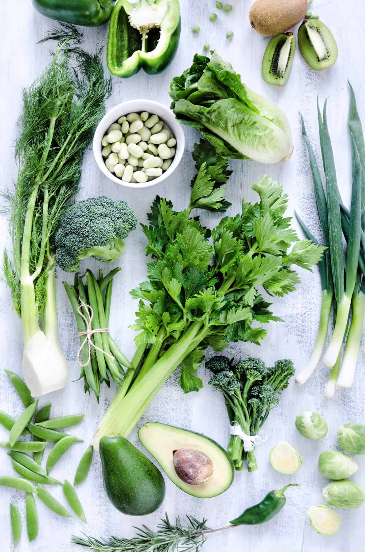 Collection of green vegetables on rustic white background from overhead, broccoli, celery, avocado, brussel sprouts, kiwi, pepper, peas, beans, lettuce,